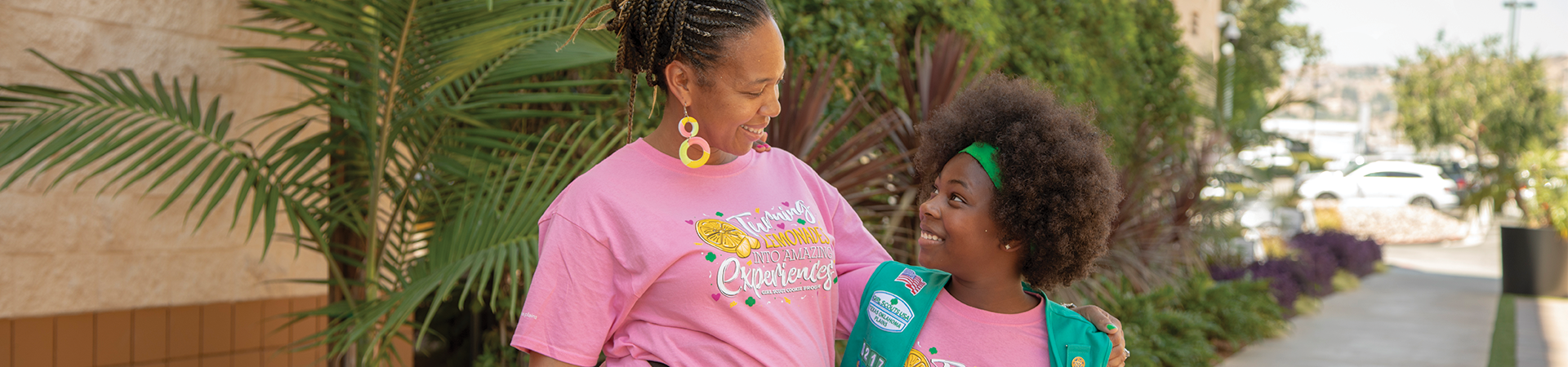  adult volunteer wearing vest with a girl scout junior (right, in baseball cap) and daisy (left) outside at a park smiling and looking at one another 